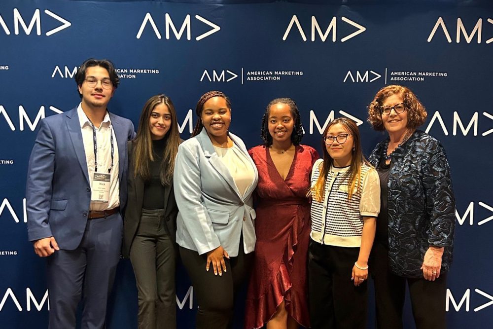 Group of five students dressed in business attire with their academic advisor posing for a photo in front of the American Marketing Association (AMA) step and repeat