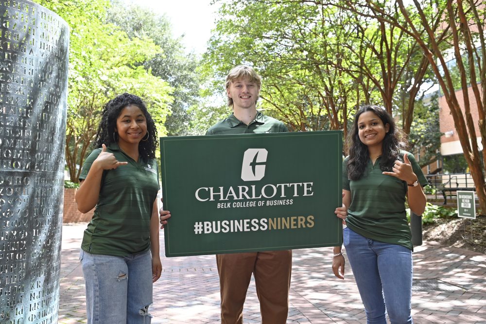 Three members of the Peer Guide program. The one on the middle is holding up a Charlotte Belk College of Business # Business Niners sign. The two on either side are making the pickaxe hand sign.