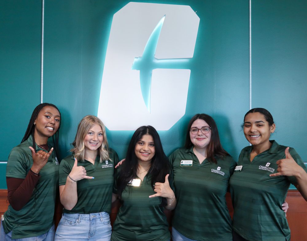 Group of five students wearing green Belk College polo shirts making the pickaxe hand sign near the all-in-c logo